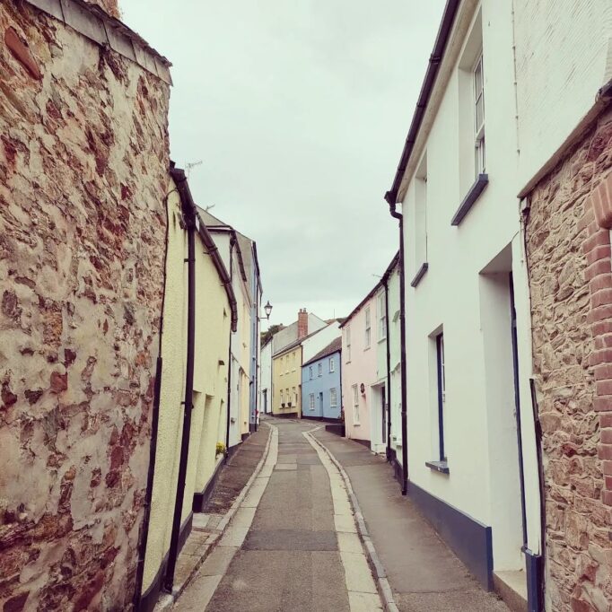 Two storey terraced houses, with natural stone and coloured render. Pale pink, yellow and blue. Narrow street in Cawsand, Cornwall with houses either side.